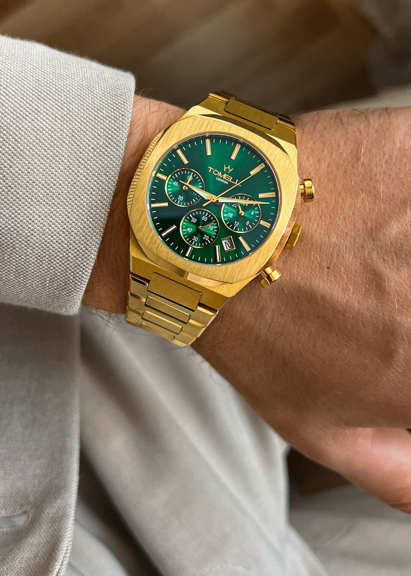 Gold watch with a green face on a wrist, wearing a light-colored shirt.