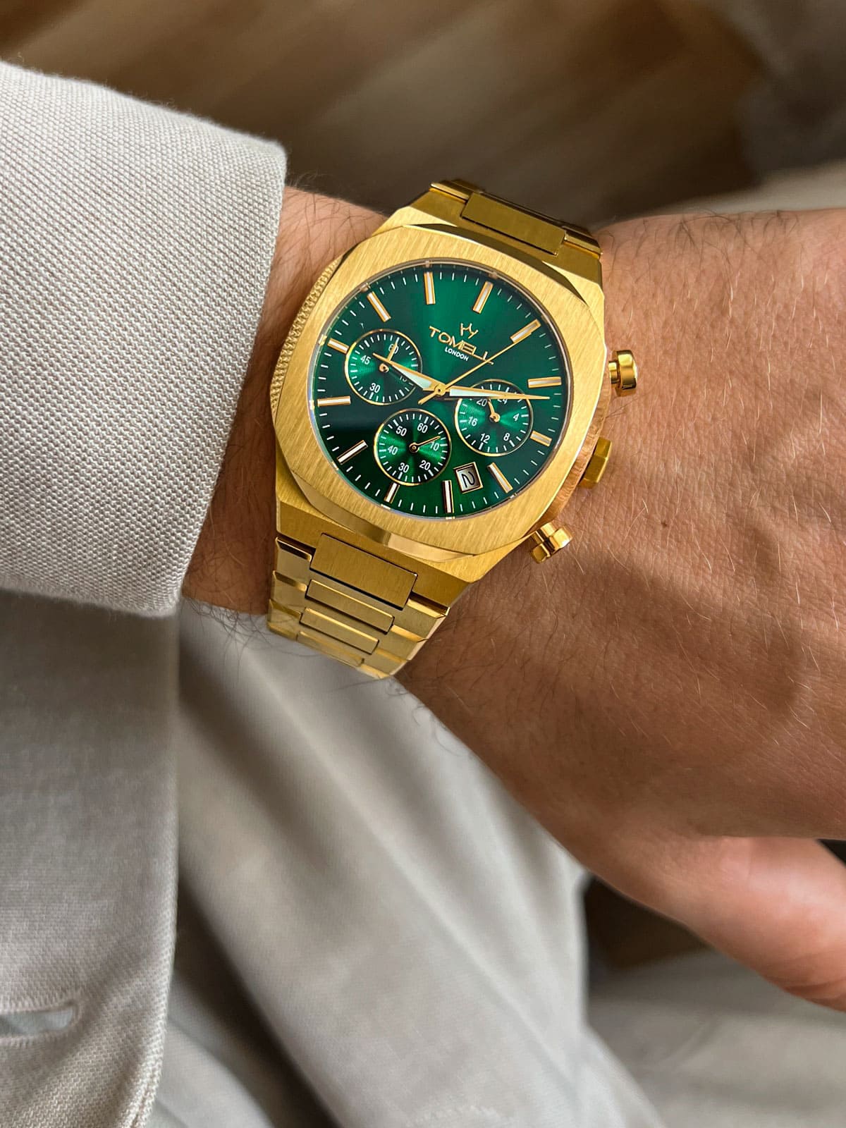 Gold watch with a green face on a wrist, wearing a light-colored shirt.